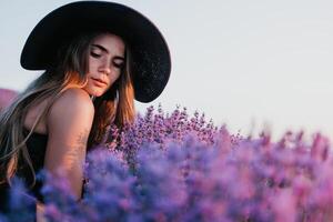 donna lavanda campo. contento spensierato donna nel nero vestito e cappello con grande orlo odorare un' fioritura lavanda su tramonto. Perfetto per ispirazione e caldo concetti nel viaggio e voglia di viaggiare. vicino su foto