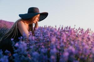 donna lavanda campo. contento spensierato donna nel nero vestito e cappello con grande orlo odorare un' fioritura lavanda su tramonto. Perfetto per ispirazione e caldo concetti nel viaggio e voglia di viaggiare. vicino su foto