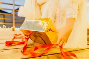 un' donna nel un' bianca vestito è Tenere un' oro scatola con un' rosso nastro. lei è indossare un' corona su sua testa. il donna appare per essere apertura il regalo scatola. foto