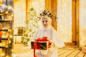 contento sorridente donna nel un' bianca vestito Tenere un' regalo scatola nel davanti di un' Natale albero. foto