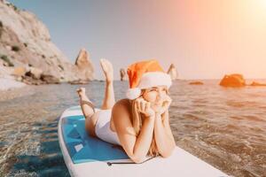 donna mare sup. vicino su ritratto di contento giovane caucasico donna con lungo capelli nel Santa cappello guardare a telecamera e sorridente. carino donna ritratto nel un' bianca bikini in posa su cenare tavola nel il mare foto