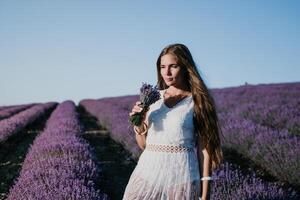 donna lavanda campo. contento spensierato donna nel un' bianca vestito a piedi nel un' lavanda campo e odorare un' lavanda mazzo su tramonto. ideale per caldo e ispirazione concetti nel voglia di girovagare e viaggio. foto