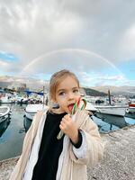 poco ragazza lecca un' lecca-lecca su un' molo contro il fondale di un' arcobaleno foto