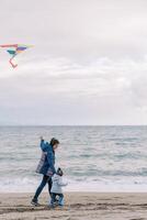 mamma con un' aquilone su un' corda passeggiate con un' poco ragazza di il mano lungo il riva del mare foto