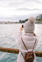 ragazza con un' zaino sta su il molo e prende immagini di il mare costa. indietro Visualizza foto