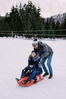 papà spinge un' sorridente mamma con un' poco ragazzo su un' slitta giù un' nevoso collina foto