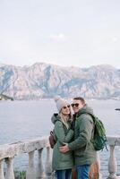 sorridente coppia abbracciare vicino il pietra balaustra al di sopra di il mare contro il fondale di montagne foto