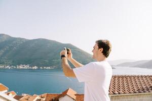 giovane uomo prende un' foto con un' smartphone di un' Visualizza di il montagne e il mare al di sopra di il rosso tetti di case