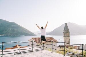 tipo salti con il suo braccia sollevato e il suo gambe piegato su il piattaforma prospiciente il baia e il campana Torre. perasto, montenegro. indietro Visualizza foto