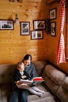 mamma legge un' libro per un' poco ragazza seduta su sua giro su un' angolo divano nel un' di legno Casa foto