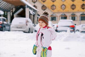 poco ragazza sta lateralmente nel il neve vicino macchine parcheggiata vicino il Hotel foto