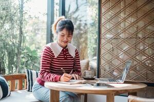 soddisfare Creatore è assorto nel scrittura Appunti nel un' taccuino Il prossimo per sua il computer portatile nel un' luminosa, naturalmente illuminato camera con un' accogliente ambiente. foto