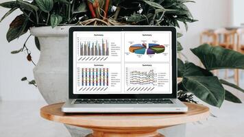 il computer portatile con dettagliato attività commerciale analitica visualizzato su schermo, posto su un' di legno tavolo vicino un' in vaso pianta. foto