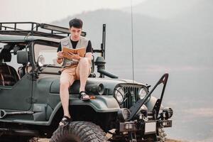 giovane uomo si siede pensieroso su il cappuccio di il suo macchina, lettura un' prenotare, con montagne nel il nebbioso distanza. foto