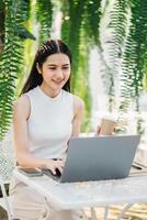 sorridente donna lavori su sua il computer portatile mentre godendo un' porta via caffè nel un' lussureggiante all'aperto bar ambientazione. foto