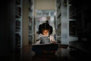 un' giovane ragazza è seduta su il pavimento nel un' biblioteca, lettura un' libro foto