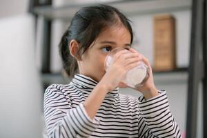 asiatico carino poco ragazza potabile latte a il tavolo a casa. gode potabile latte. foto