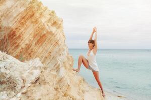 un' bellissimo giovane ragazza con corto capelli è vestito nel pantaloncini e un' bianca maglia praticante yoga contro il fondale di il mare e montagne. posa guerriero eroe. concetto di equilibrio e quiete foto