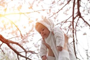 contento famiglia. madre mani lanci su bambino nel il fioritura Mela alberi, su soleggiato giorno nel il parco. positivo umano emozioni, sentimenti. ciliegia e Mela fiore. primavera volta. foto