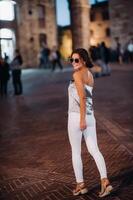 un' ragazza nel i pantaloni e un' maglietta con bicchieri nel il notte città di san gimignano.a ragazza passeggiate in giro il città nel italia.toscana. foto