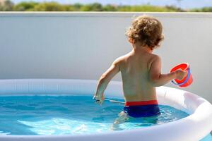 indietro Visualizza di un' poco ragazzo pesca con un' netto nel il balcone piscina acqua su un' caldo estate vacanza giorno. lui è Tenere un' rosso benna. Visualizza di cristallo pulito acqua superficie, sole brillare luminosa piscina. foto