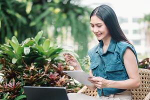 giovane donna Tenere credito carta e utilizzando ltablet. un' sorridente giovane donna impegna con un' tavoletta in mezzo vivace verde fogliame. in linea acquisti, commercio elettronico, Internet bancario, la spesa i soldi. foto