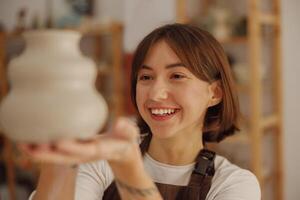 sorridente femmina vasaio indossare grembiule Tenere non sparato argilla vaso mentre in piedi nel ceramica studio foto