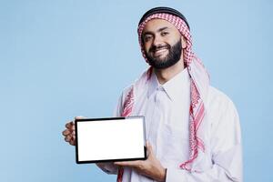 sorridente arabo uomo presentazione digitale tavoletta vuoto schermo per App presentazione e guardare a telecamera con allegro espressione. arabo nel tradizionale Abiti mostrando vuoto touch screen nel orizzontale modalità foto