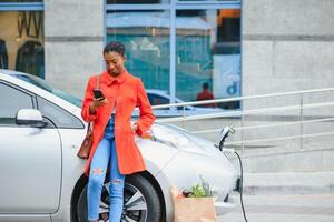 ricarica elettro auto a il elettrico gas stazione. africano americano ragazza in piedi di il auto foto