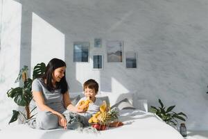 mamma con figlio nel pigiama mangiare frutta su letto. ritratto felice, sorridente, gioioso famiglia, madre e figlio avendo prima colazione nel letto, sorpresa su mamma giorno. positivo umano emozioni foto