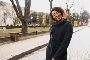 attraente contento di carnagione scura modello con afro acconciatura e piercing al naso, in posa all'aperto contro urbano sfondo durante sua mattina camminare, guardare giù con timido Sorridi mostrando sua bianca denti. bagliore sole foto