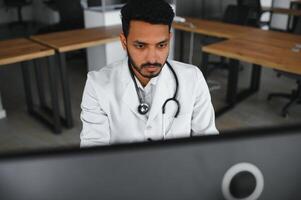 indiano maschio medico medico indossare occhiali, bianca medico toga e stetoscopio seduta a il scrivania con il il computer portatile nel moderno clinica e coinvolti in linea video conferenza, consulenza a distanza. foto