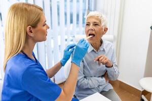 anziano paziente apertura sua bocca per il medico per Guarda nel sua gola. femmina medico l'esame dolorante gola di paziente nel clinica. otorinolaringoiatra esamina dolorante gola di paziente. foto