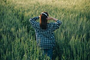 giovane bella donna nel rosso estate vestito e cannuccia cappello a piedi su giallo azienda agricola campo con maturo d'oro Grano godendo caldo sera. foto