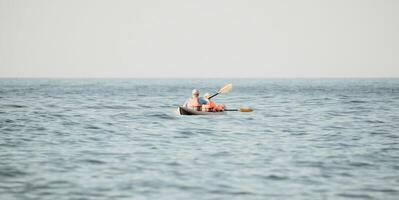 contento coppia kayak nel un gonfiabile kayak su il mare a tramonto. coppia canoa nel il mare vicino il isola con montagne. persone kayak nel vita giacche vela. indietro Visualizza foto