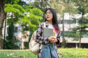 un' giovane asiatico femmina Università alunno trasporto un' zaino e roba, in piedi nel il città universitaria parco. foto