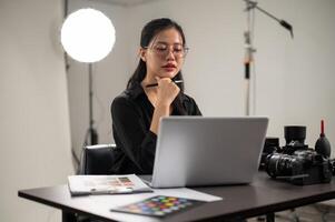 un' fiducioso asiatico femmina fotografo è Lavorando su sua computer portatile, Lavorando nel un' servizio fotografico studio. foto