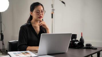 un' fiducioso asiatico femmina fotografo è Lavorando su sua computer portatile, Lavorando nel un' servizio fotografico studio. foto