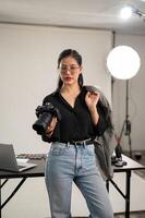 un' fiducioso asiatico femmina fotografo è in piedi nel sua studio con un' dslr telecamera nel sua mano. foto
