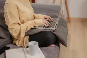 un' donna nel comodo Abiti Lavorando su sua il computer portatile computer su il divano nel il vivente camera. foto