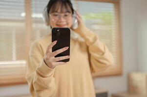 un' carino asiatico donna è ascoltando per musica su sua cuffie e utilizzando sua Telefono nel il vivente camera. foto