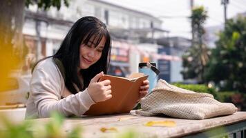 un attraente, contento asiatico donna è lettura un' libro a un' tavolo all'aperto nel il città. foto