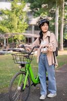 un' giovane asiatico femmina Università alunno indossare un' bicicletta casco è spingendo sua bicicletta nel un' verde parco. foto