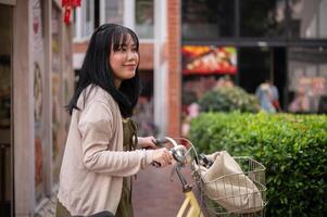un' Bellissima, positivo asiatico donna nel un' carino vestito è a piedi e spingendo sua bicicletta nel il città. foto