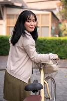 un' sorridente, contento giovane asiatico donna a piedi e spingendo sua bicicletta nel il città. foto
