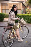 un' sorridente, contento giovane asiatico donna nel un' carino vestito è equitazione un' bicicletta nel il città foto