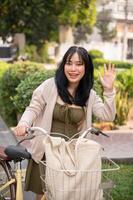 un' amichevole asiatico donna onde sua mano per dire Ciao per sua amico mentre spingendo sua bicicletta nel il città. foto