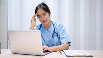 un' stressato, oberati di lavoro, e stanco asiatico femmina medico è lettura sua medico casi su un' il computer portatile. foto