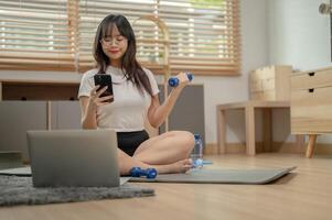 un' contento asiatico donna è esercizio con un' manubrio mentre chat con sua amici su sua smartphone. foto