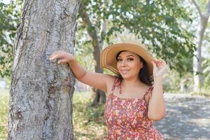 donna indossare cappello e vestito con fiori ammirazione natura nel un' pubblico parco vicino per un' albero. soleggiato estate giorno. foto